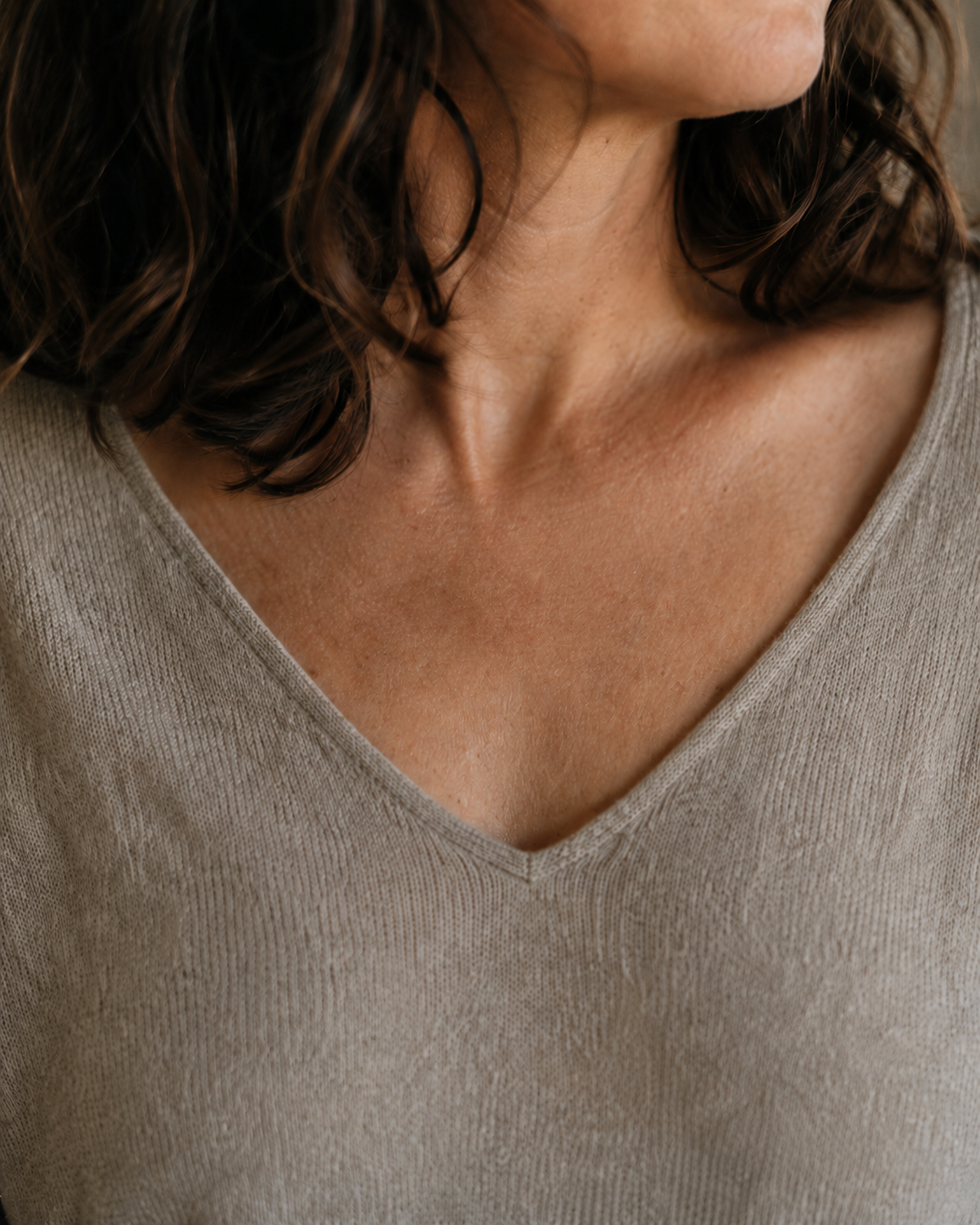 A close, warm photograph — a woman's collarbone and the base of her turned neck above a soft linen V-neck, where a pendant would rest
