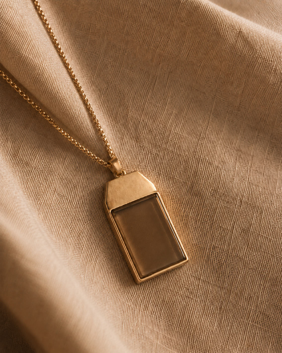 The Somatag pendant — a brushed-gold tag with a tiger's-eye stone face — resting on warm linen in golden light